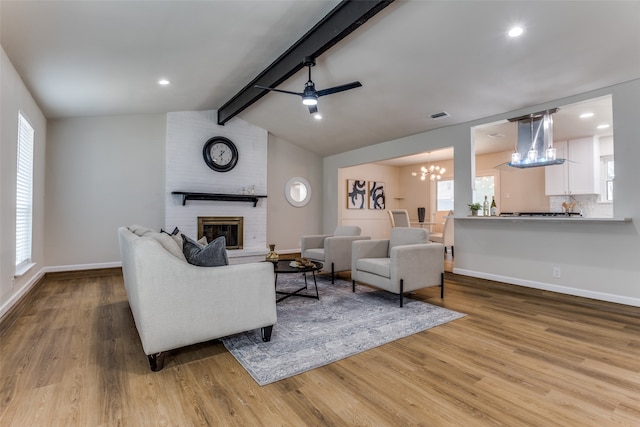 living room with vaulted ceiling with beams, light hardwood / wood-style flooring, ceiling fan with notable chandelier, and a brick fireplace