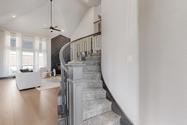 stairs with a fireplace, ceiling fan, hardwood / wood-style floors, and high vaulted ceiling