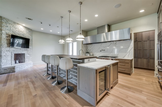 kitchen with tasteful backsplash, wall chimney range hood, a fireplace, a center island, and light hardwood / wood-style floors