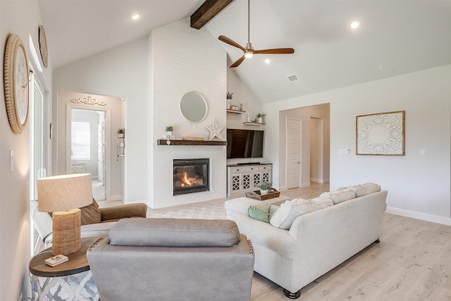 living room featuring high vaulted ceiling, ceiling fan, light wood-type flooring, beam ceiling, and a large fireplace