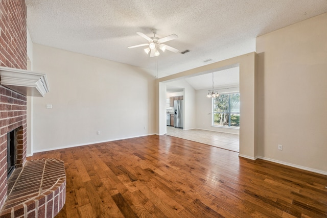 unfurnished living room with a textured ceiling, ceiling fan with notable chandelier, hardwood / wood-style flooring, and a brick fireplace