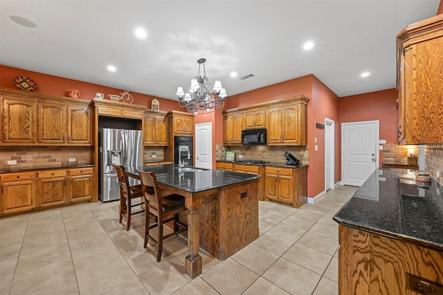 kitchen with black microwave, brown cabinetry, an island with sink, stainless steel fridge, and decorative light fixtures