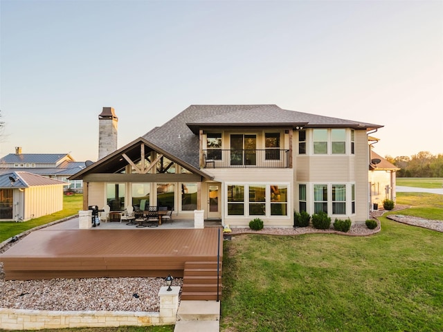 back of property at dusk featuring roof with shingles, a deck, a chimney, a balcony, and a yard