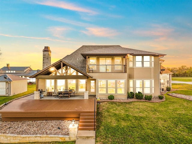 back house at dusk with a wooden deck, a balcony, a lawn, and a storage shed