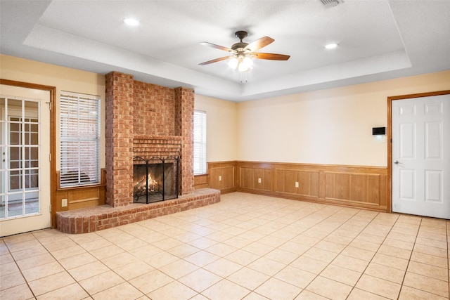 unfurnished living room with a brick fireplace, ceiling fan, light tile patterned floors, and a tray ceiling