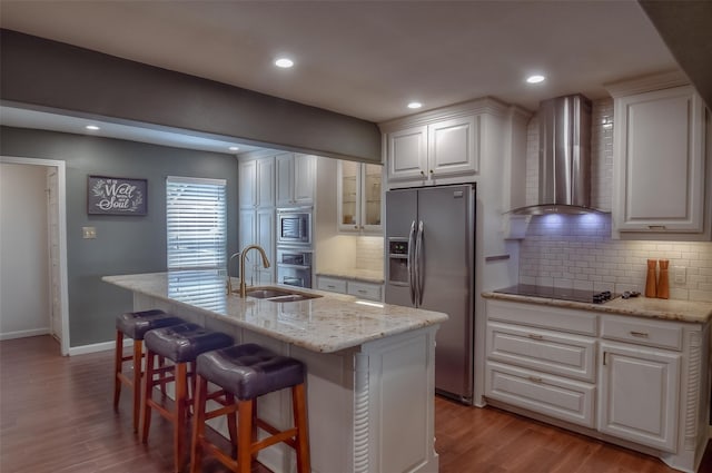 kitchen with stainless steel appliances, wall chimney range hood, an island with sink, light hardwood / wood-style floors, and white cabinets