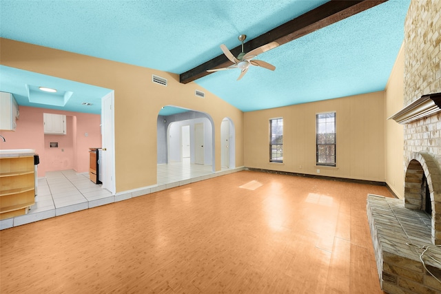 unfurnished living room featuring a brick fireplace, a textured ceiling, ceiling fan, lofted ceiling with beams, and light hardwood / wood-style floors