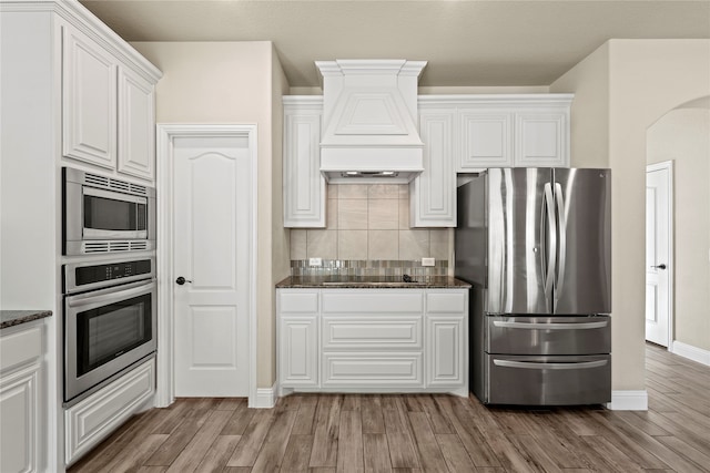 kitchen with white cabinetry, stainless steel appliances, premium range hood, backsplash, and light hardwood / wood-style floors