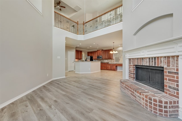 unfurnished living room featuring ceiling fan with notable chandelier, light hardwood / wood-style floors, a high ceiling, and a brick fireplace