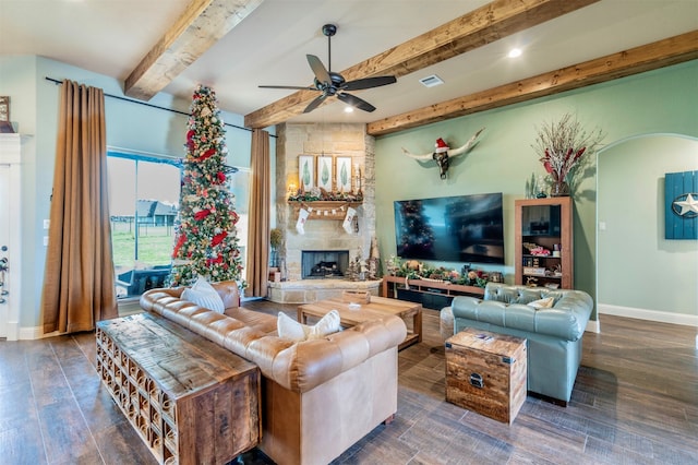 living room featuring a stone fireplace, ceiling fan, beamed ceiling, and dark wood-type flooring