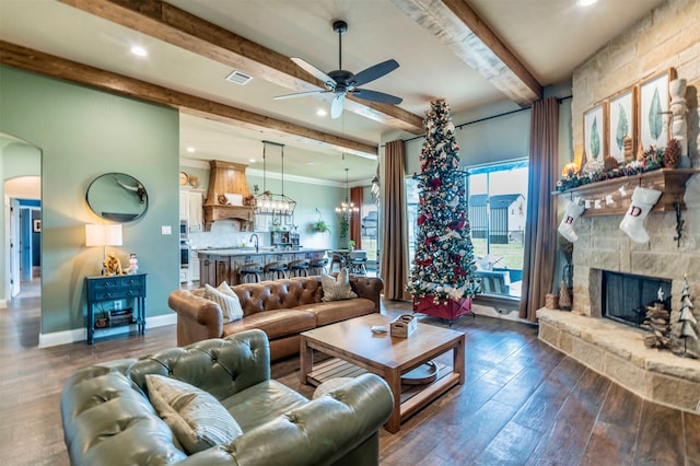 living room featuring dark wood-type flooring, ceiling fan with notable chandelier, a stone fireplace, sink, and beam ceiling