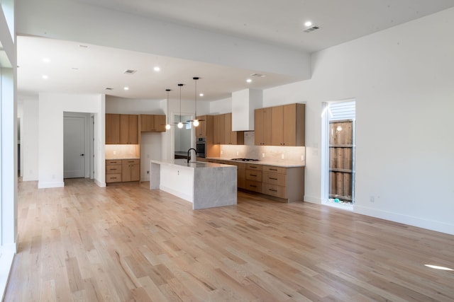 kitchen with tasteful backsplash, decorative light fixtures, light hardwood / wood-style floors, oven, and an island with sink