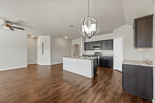 kitchen featuring dark hardwood / wood-style flooring, ceiling fan with notable chandelier, stainless steel appliances, decorative light fixtures, and a center island with sink