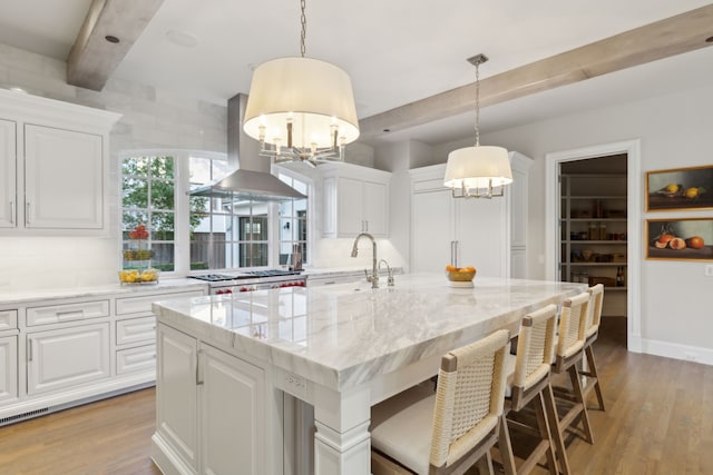 kitchen featuring light stone countertops, island range hood, a kitchen island with sink, white cabinetry, and hanging light fixtures