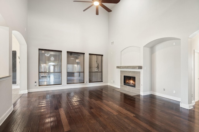 unfurnished living room with a tile fireplace, ceiling fan, a towering ceiling, and dark wood-type flooring