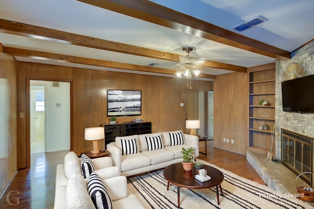 living room featuring wood walls, ceiling fan, light wood-type flooring, a fireplace, and beamed ceiling