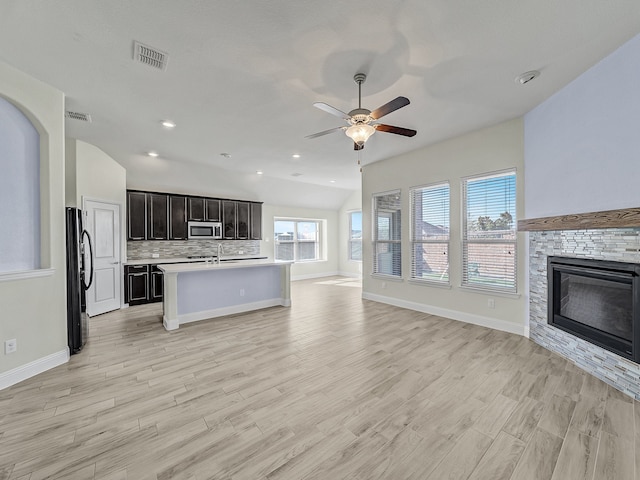 kitchen with ceiling fan, stainless steel appliances, a stone fireplace, a kitchen island with sink, and light wood-type flooring