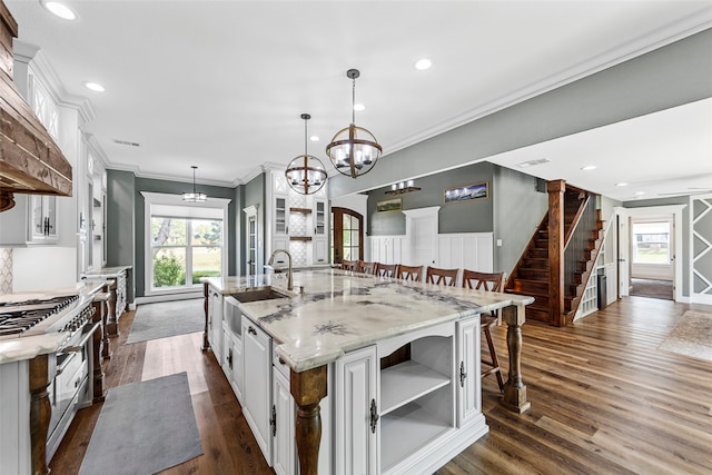 kitchen with ornamental molding, light stone countertops, white cabinets, a center island with sink, and decorative light fixtures