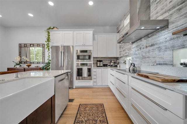 kitchen with white cabinetry, wall chimney range hood, light stone counters, light hardwood / wood-style flooring, and appliances with stainless steel finishes