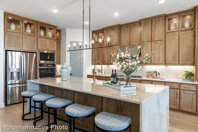 kitchen with appliances with stainless steel finishes, tasteful backsplash, light stone counters, a kitchen island with sink, and light hardwood / wood-style floors