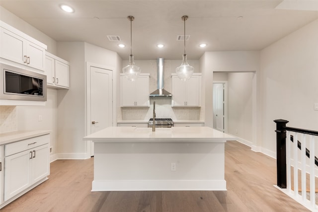 kitchen featuring wall chimney range hood, decorative light fixtures, white cabinets, and an island with sink