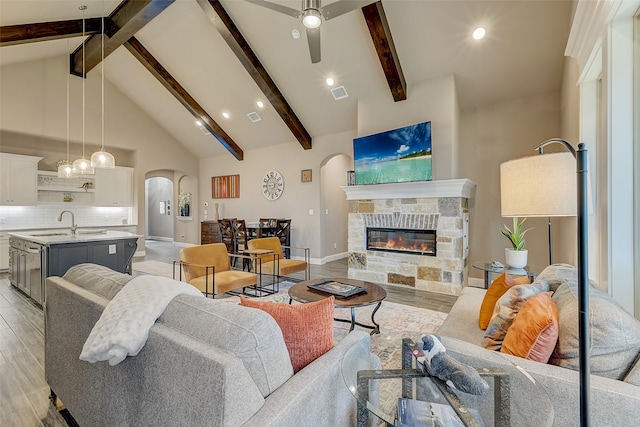 living room with ceiling fan, beam ceiling, high vaulted ceiling, light hardwood / wood-style flooring, and a stone fireplace