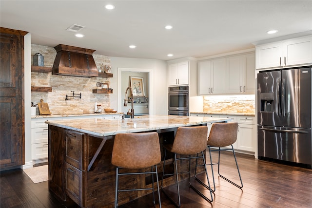 kitchen with light stone counters, stainless steel appliances, a kitchen island with sink, dark wood-type flooring, and white cabinetry