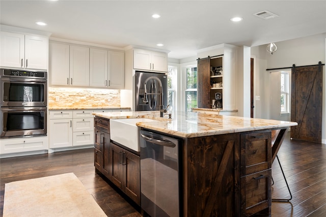 kitchen featuring a kitchen island with sink, white cabinets, dark hardwood / wood-style floors, a barn door, and stainless steel appliances