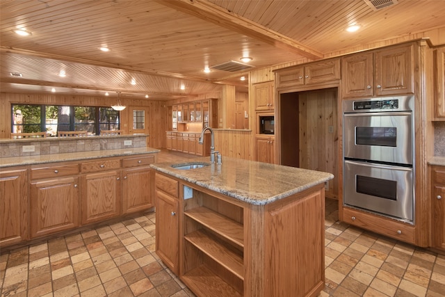 kitchen with wood ceiling, stainless steel double oven, sink, a center island with sink, and wood walls