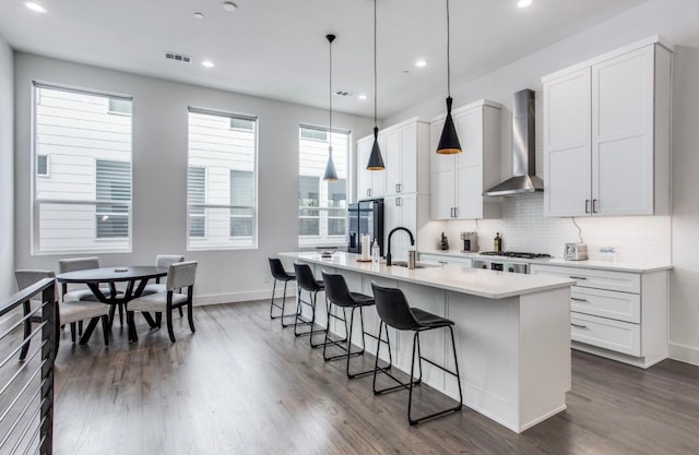 kitchen with sink, wall chimney range hood, decorative light fixtures, white cabinets, and an island with sink