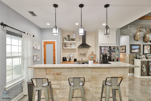 kitchen with white cabinetry, wall chimney exhaust hood, black fridge, a kitchen bar, and decorative backsplash