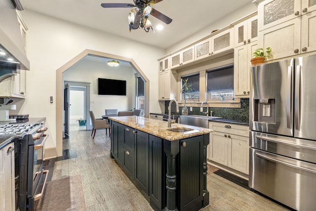 kitchen with arched walkways, a kitchen island with sink, a sink, appliances with stainless steel finishes, and light wood finished floors