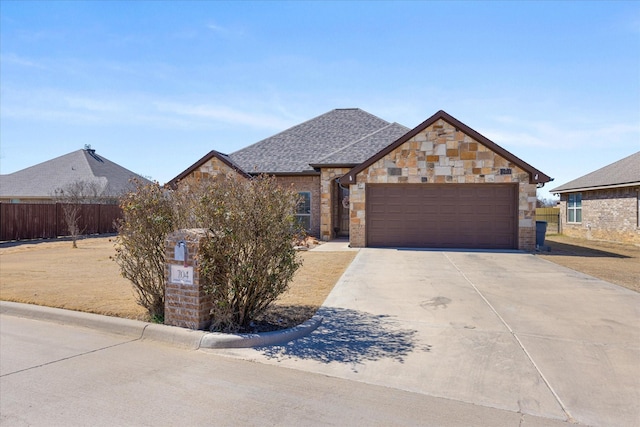 french country inspired facade featuring a shingled roof, concrete driveway, an attached garage, fence, and stone siding