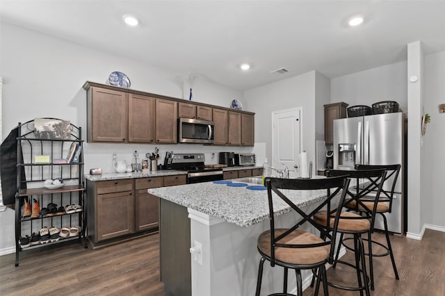 kitchen featuring dark brown cabinetry, dark wood-type flooring, light stone counters, a center island with sink, and appliances with stainless steel finishes