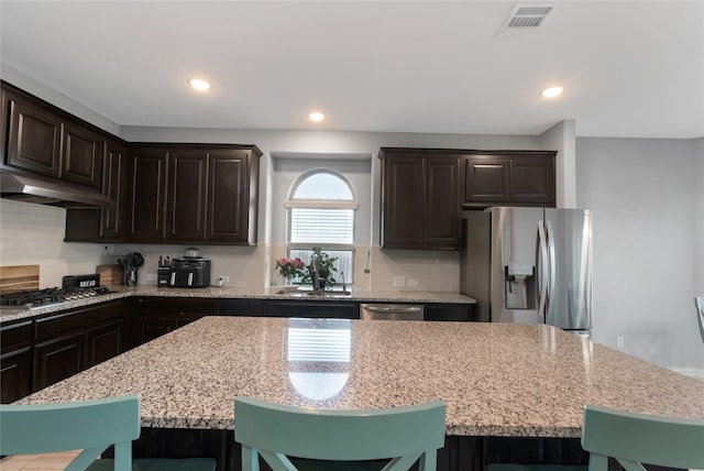 kitchen featuring appliances with stainless steel finishes, light stone counters, dark brown cabinetry, a kitchen island, and a breakfast bar area