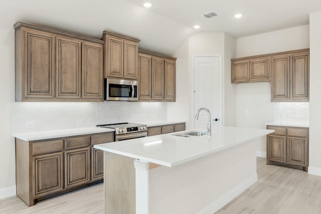 kitchen with decorative backsplash, light wood-type flooring, stainless steel appliances, sink, and an island with sink