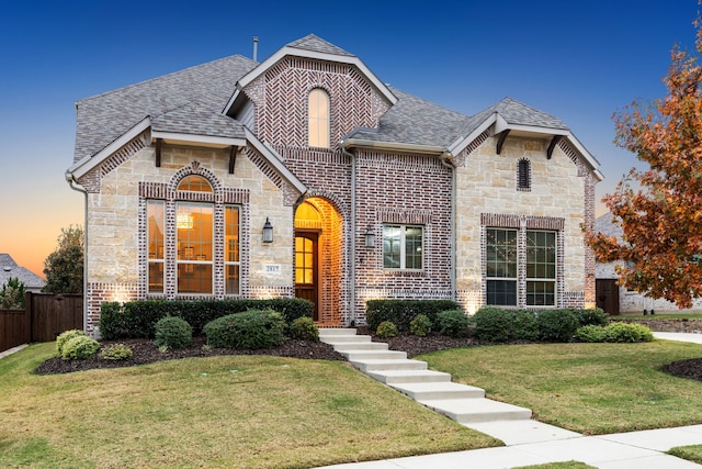 french country style house featuring brick siding, a shingled roof, fence, stone siding, and a lawn