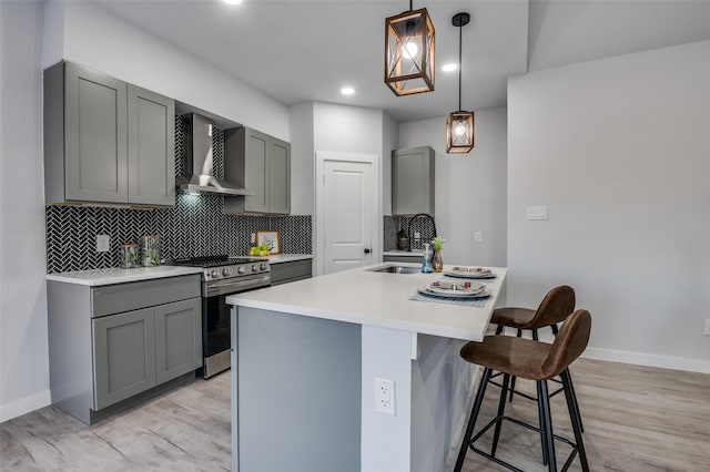 kitchen with gray cabinetry, a center island with sink, a kitchen breakfast bar, stainless steel stove, and wall chimney exhaust hood