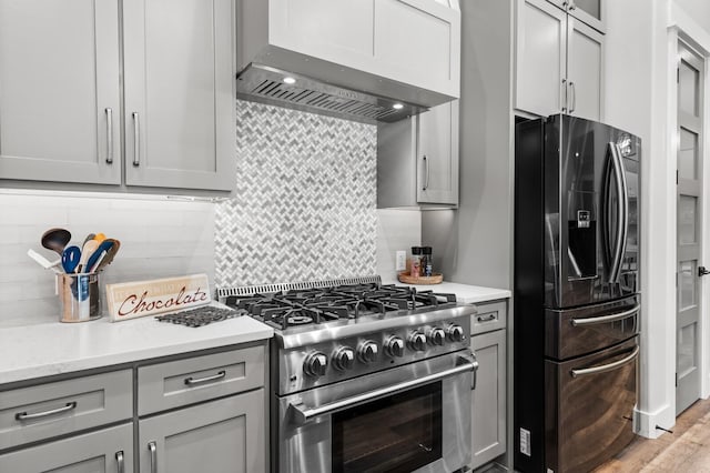 kitchen featuring gray cabinets, backsplash, light stone counters, stainless steel appliances, and wall chimney range hood