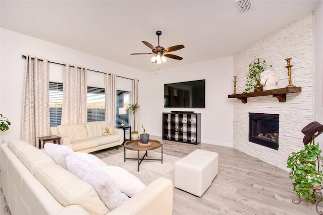 living room featuring visible vents, light wood-style floors, ceiling fan, a stone fireplace, and baseboards