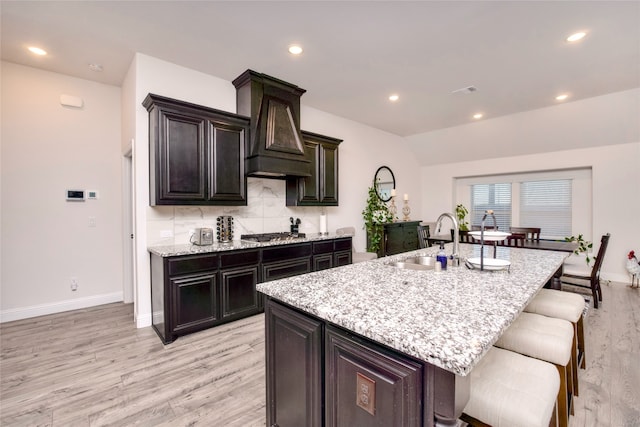 kitchen with sink, light hardwood / wood-style flooring, an island with sink, dark brown cabinets, and custom range hood