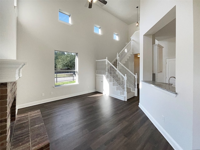 unfurnished living room with a brick fireplace, ceiling fan, dark wood-type flooring, sink, and a high ceiling