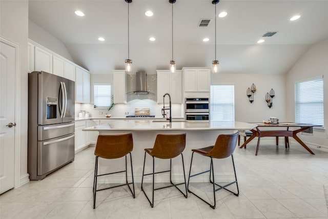 kitchen featuring appliances with stainless steel finishes, a center island with sink, vaulted ceiling, and wall chimney exhaust hood