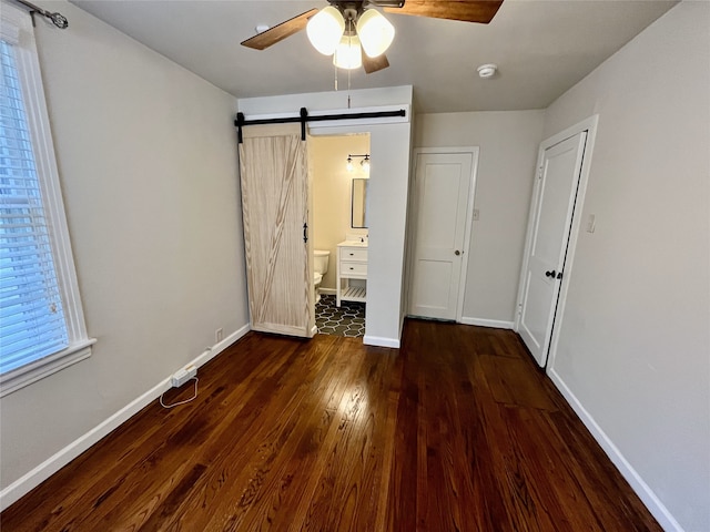 unfurnished bedroom with a barn door, dark hardwood / wood-style floors, ensuite bath, and ceiling fan