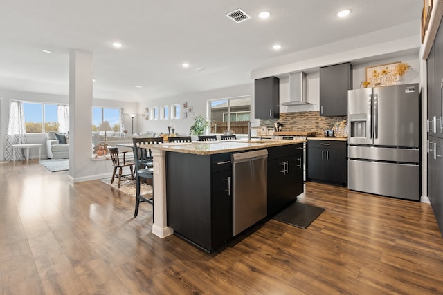 kitchen featuring a center island with sink, wall chimney range hood, light stone counters, wood-type flooring, and stainless steel appliances