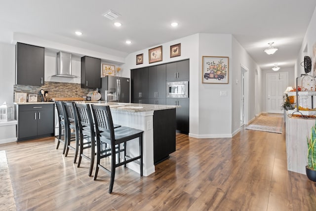 kitchen with appliances with stainless steel finishes, light wood-type flooring, a breakfast bar, wall chimney range hood, and an island with sink