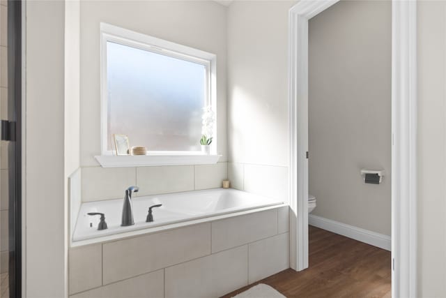 bathroom with a relaxing tiled tub, toilet, and wood-type flooring