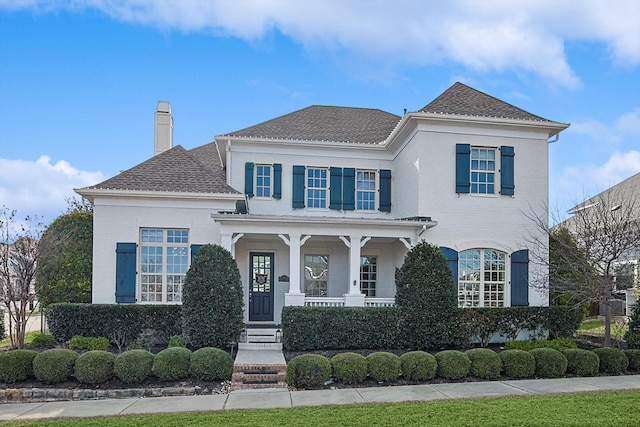 view of front of house featuring covered porch, a shingled roof, a chimney, and brick siding