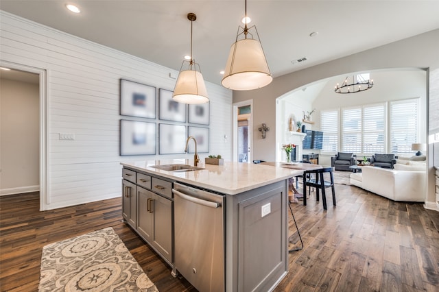 kitchen featuring dishwasher, dark wood-type flooring, hanging light fixtures, light stone counters, and an island with sink