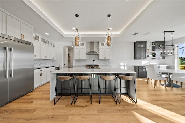 kitchen with built in refrigerator, a kitchen island with sink, wall chimney range hood, and white cabinets